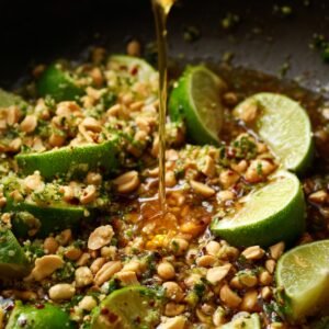 A close-up of fresh lime wedges, crushed peanuts, and herbs being drizzled with sauce, capturing the finishing flavors of Pad Thai.