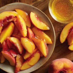 Bowl of fresh peach wedges lightly coated in honey, with additional peaches and a honey jar on a wooden table.