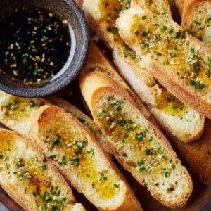 Golden toasted baguette slices brushed with olive oil and herbs, served next to a small bowl of herb oil dip.