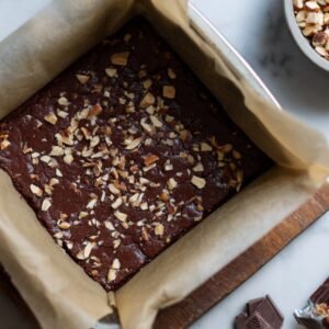 A pan of baked brownies topped with chopped nuts, set on parchment paper with ingredients nearby.