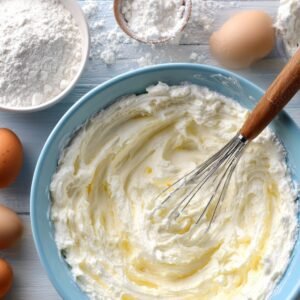 A mixing bowl filled with smooth cream cheese mixture, surrounded by flour, sugar, and eggs on a light blue table.