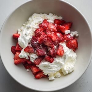 Mixing bowl with whipped cream cheese, sugar, and diced strawberries coated in sugar, forming the filling mixture.