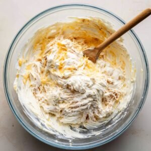 A glass mixing bowl with a wooden spoon stirring together cream cheese, shredded chicken, and cheese, forming the buffalo chicken dip mixture.