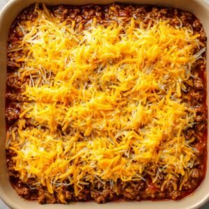 A baking dish filled with buffalo chicken dip topped with a generous layer of shredded cheddar and Monterey Jack cheese before baking.