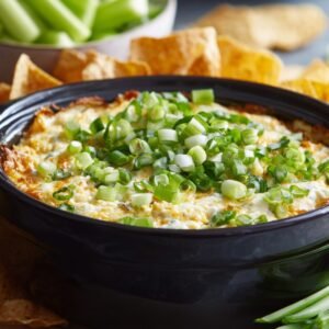 A bowl of creamy buffalo chicken dip topped with chopped green onions, surrounded by tortilla chips and celery sticks for dipping.