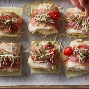 Raw chicken rollatini wrapped with prosciutto, sprinkled with shredded cheese and herbs, ready to bake.