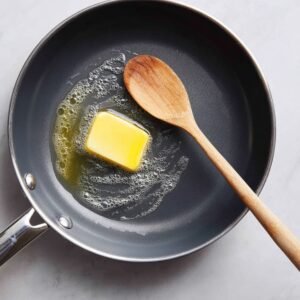 "Non-stick skillet with a pat of butter melting and a wooden spoon, preparing the pan for making crepes."