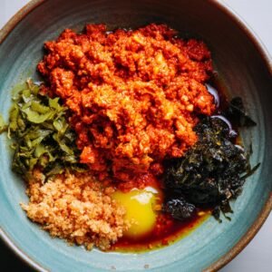 A bowl filled with various ingredients for making kimchi, including bright red chili paste, a whole egg, seaweed, and fermented rice. The ingredients are arranged neatly in the bowl, ready to be combined for the kimchi preparation.