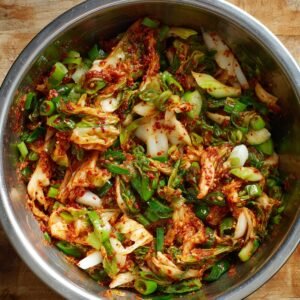 A large stainless steel mixing bowl filled with chopped napa cabbage, green onions, and a vibrant red chili paste, all mixed together. The kimchi ingredients are coated in a spicy red sauce, indicating it's freshly made and packed with flavor.