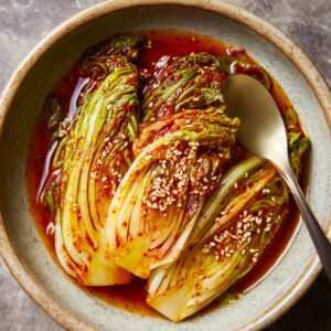 A bowl of fresh kimchi with vibrant red chili paste, sesame seeds, and crisp napa cabbage. The kimchi is coated in a spicy, tangy marinade with a shiny, glossy texture, ready to be served.