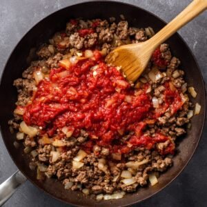 A skillet containing browned ground beef and onions, with tomato paste being stirred in with a wooden spoon.