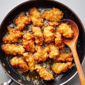 Pieces of fried chicken coated in a sticky orange sauce cooking in a pan with a wooden spoon.