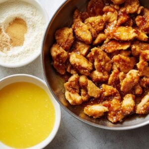 A bowl of crispy chicken pieces coated in orange sauce, with bowls of flour, seasoning, and beaten egg beside it.