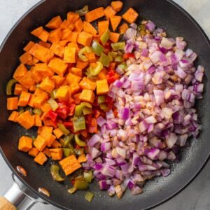 Uncooked sweet potato cubes, green bell peppers, red peppers, and diced red onions arranged in a skillet before cooking.