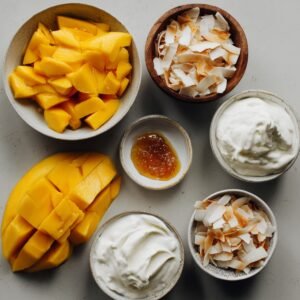 "Flat lay of ingredients for mango pancakes including flour, eggs, butter, sugar, yogurt, and ripe mango pieces."