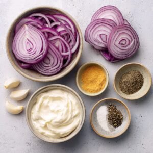 Fresh ingredients for cucumber salad including sliced red onions, garlic cloves, mayonnaise, black pepper, salt, and seasonings in small bowls.