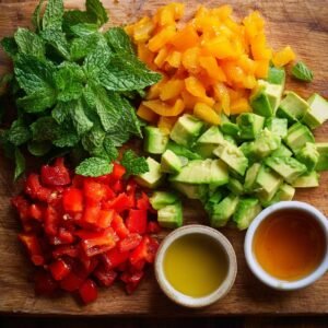 A rustic wooden board with piles of diced avocado, yellow peppers, red peppers, fresh mint leaves, and small bowls of olive oil and honey.