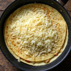 Tortilla in a pan topped generously with shredded cheese, ready to be folded and cooked into a quesadilla.