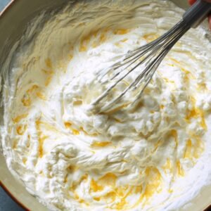A bowl of whipped egg whites being gently folded into a yellow corn mixture using a metal whisk, creating a fluffy texture.