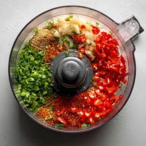A food processor filled with fresh ingredients: chopped red chili peppers, green herbs, onions, garlic, and various spices, ready to be blended into a sauce or paste.