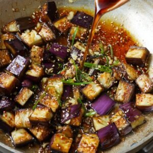Cubes of Japanese eggplant being cooked in a skillet, drizzled with a glossy soy-based sauce, sprinkled with sesame seeds, garlic, and scallions.