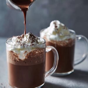 A glass mug of rich hot chocolate topped with whipped cream and chocolate shavings, while more chocolate sauce is being poured on top. Another mug of hot chocolate sits in the background.