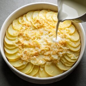 Sliced potatoes arranged in baking dish with diced onions and shredded cheese, milk being poured over.