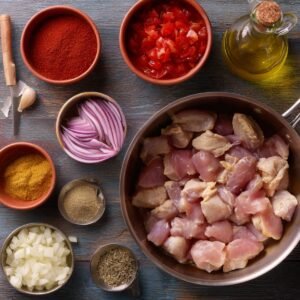 Raw diced chicken pieces in a bowl surrounded by spices, chopped onions, sliced red onions, tomatoes, garlic, and olive oil, laid out for cooking.