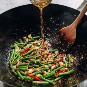 A wok with green beans being stir-fried with garlic, red chili, and spices. The cook is pouring oil into the pan, adding a shiny golden liquid to the mix.