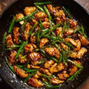 A close-up shot of Black Pepper Chicken, featuring tender pieces of chicken cooked in a dark savory sauce with green onions. The dish is served in a wok with a glossy texture from the sauce.