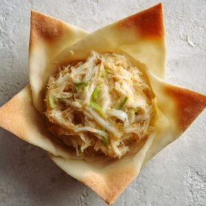 A close-up of a single Rangoon crab bomb sitting on a surface with a filling of shredded crab, cabbage, and green onions inside a crispy, golden wrapper.