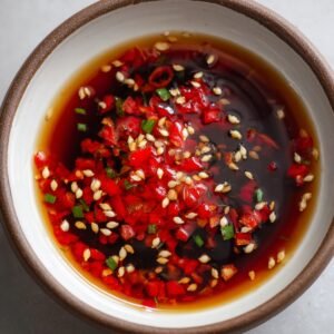 A small bowl filled with soy sauce, chopped red chilies, and sesame seeds, garnished with sliced green onions, creating a spicy dipping sauce.